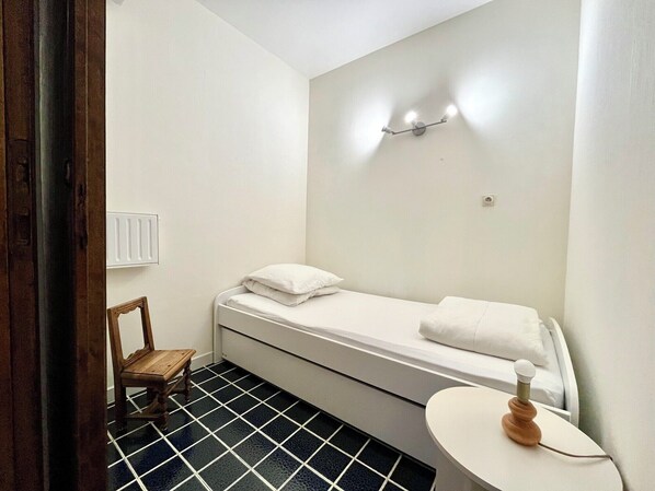 A small, clean room with a single bed, a wooden chair, a small table, and a wall-mounted light fixture.