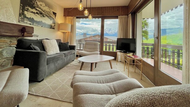 A living room with a sofa, armchairs, a coffee table, and a TV. There is a large window with a view of mountains. - 80 m² apartment with terrace (Combloux)