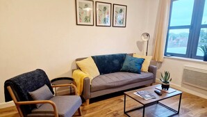 Un salon avec un canapé gris, un fauteuil en bois, une table basse et un lampadaire. Trois tableaux encadrés ornent le mur.