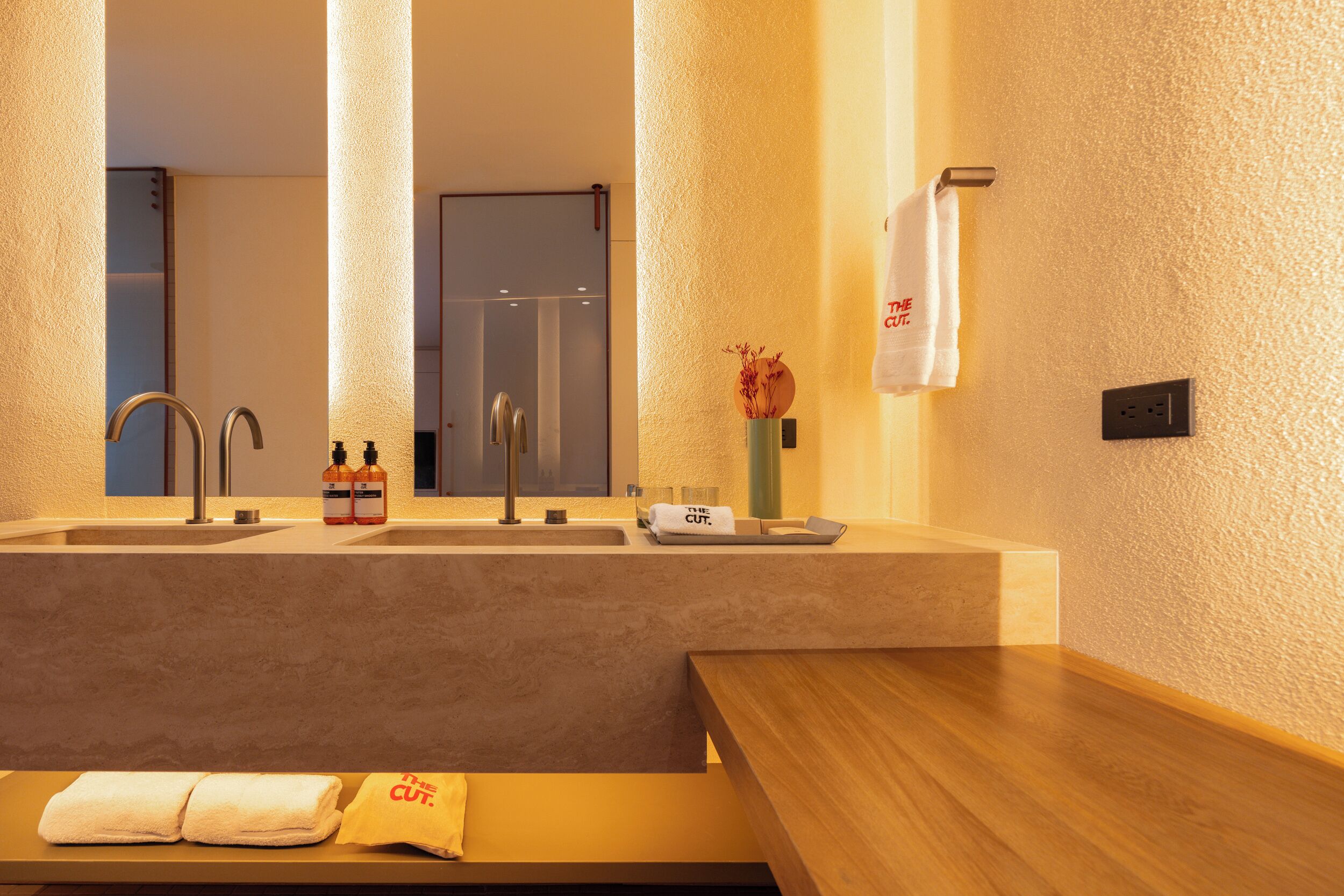 Een moderne badkamer met een grote spiegel, twee wasbakken, een bankje met handdoeken en een vaas met gedroogde bloemen.