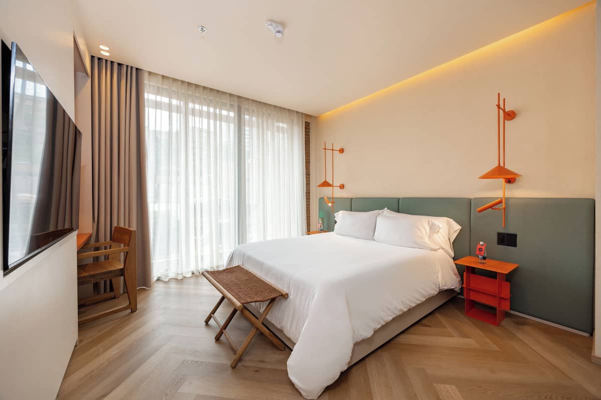 Quarto de hotel moderno com uma cama grande, um banco de madeira, uma televisão e uma escrivaninha com abajur.