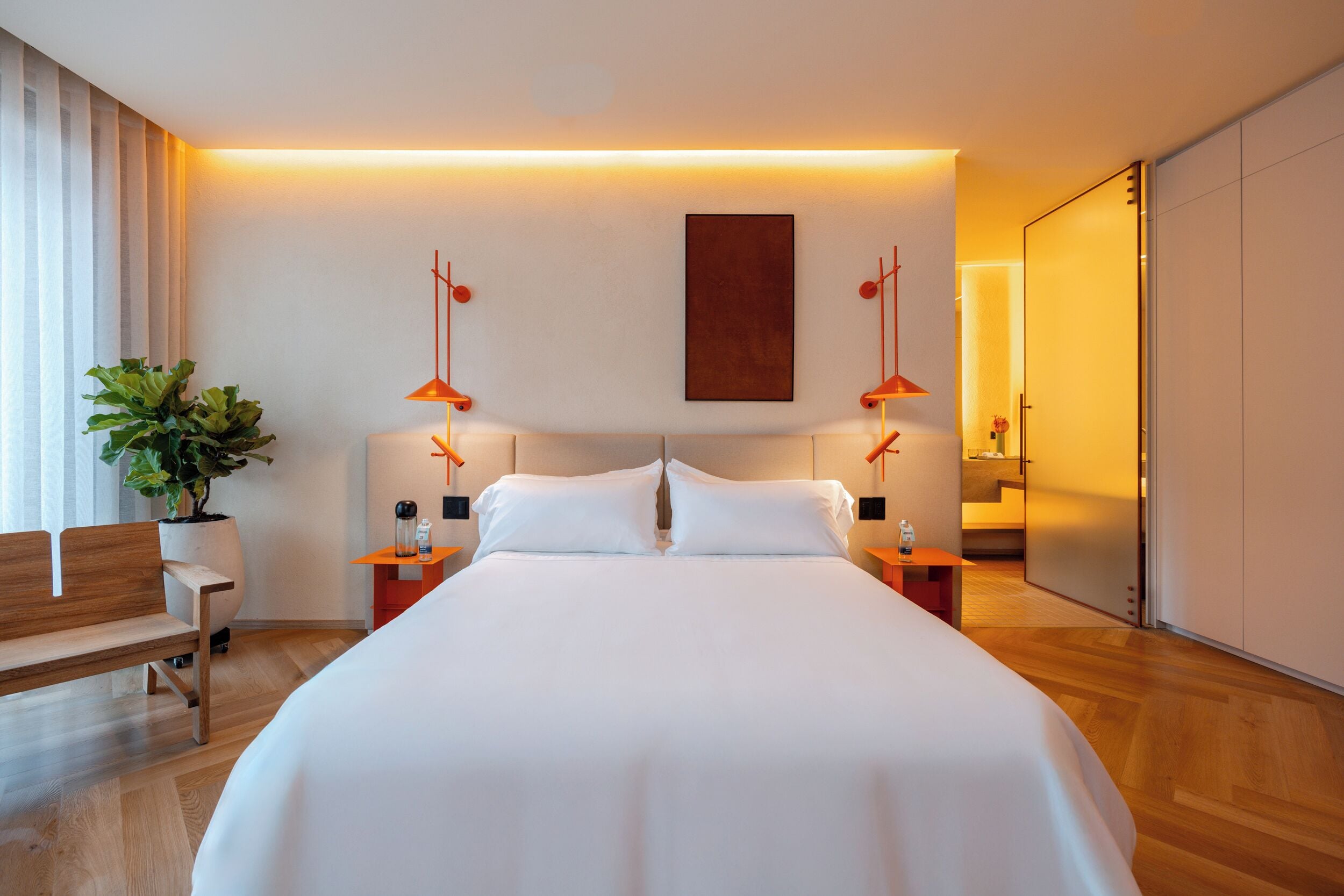 A modern hotel room with a large bed, bedside tables, a bench, and a potted plant.
