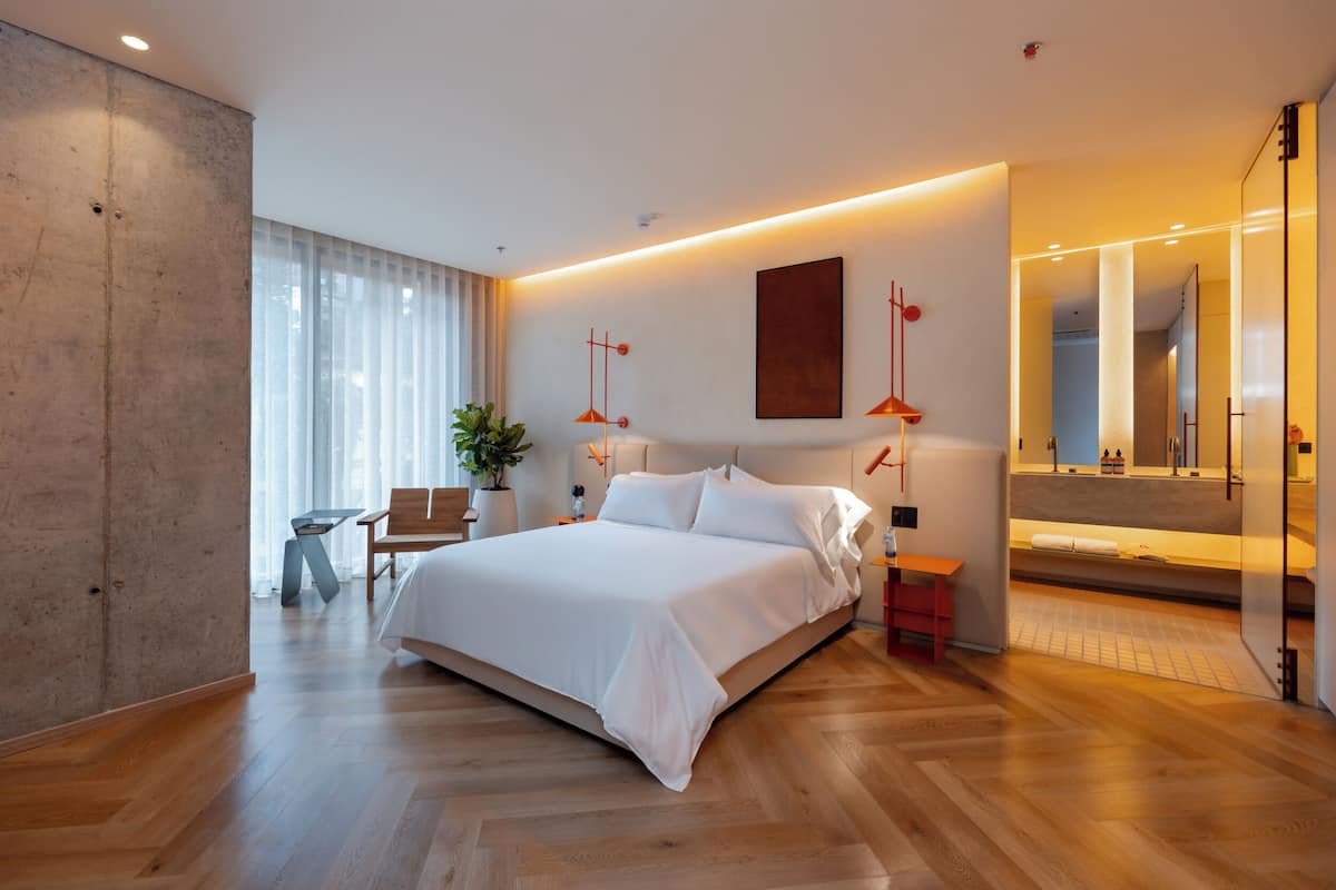 Quarto moderno com uma cama grande, mesas de cabeceira, um espaço de estar com uma poltrona e mesa, e um espelho amplo.