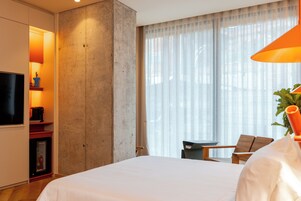 A hotel room with a large bed, a small table, a chair, a TV, and a plant.