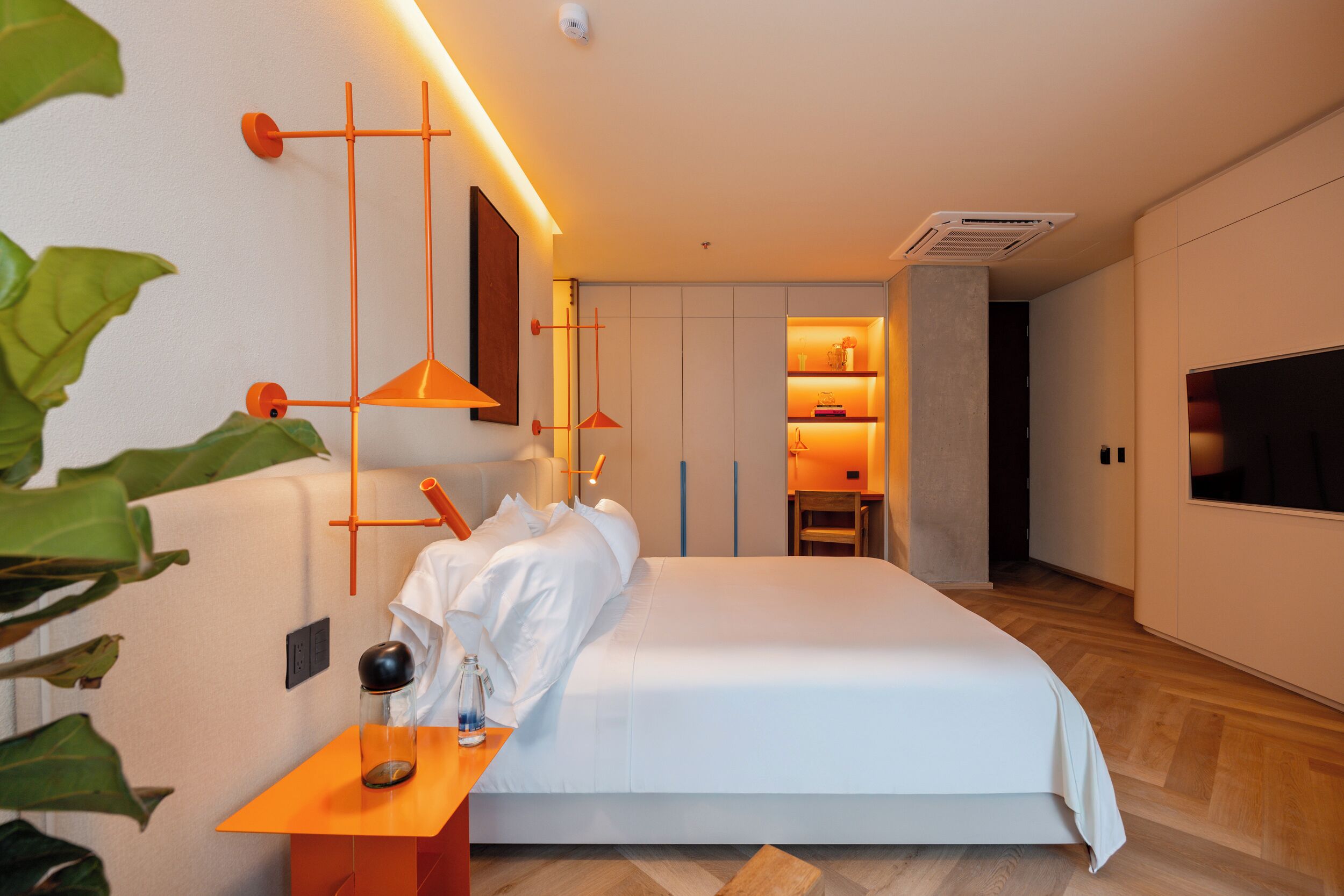 A modern hotel room with a large bed, a bedside table with a lamp, a TV, and a small seating area with a chair and table.