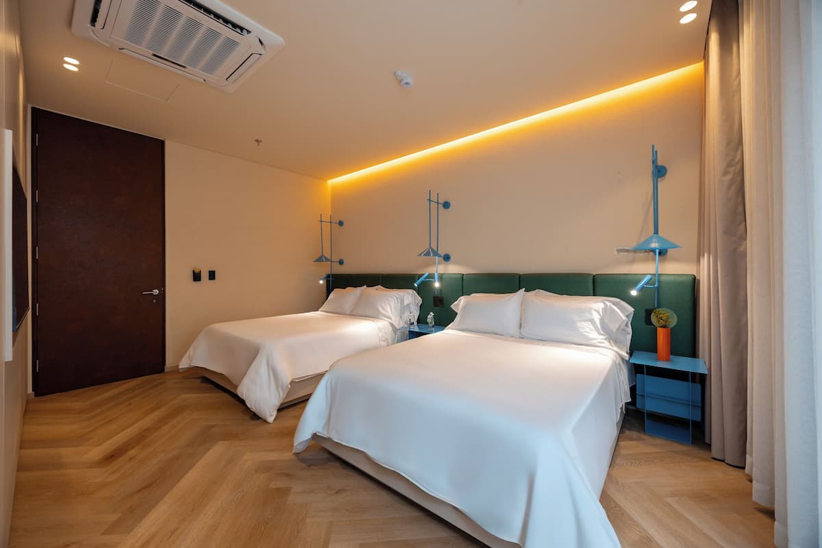 Quarto de hotel com duas camas, piso de madeira, ar-condicionado no teto e mesa de cabeceira com abajur.