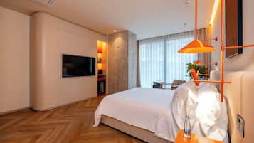 Una habitación de hotel moderna con una cama grande, un televisor de pantalla plana, una mesita con un florero y un ventanal con cortinas.