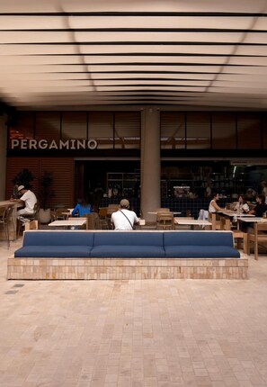 Un área de estar al aire libre con mesas y bancos, una pérgola encima y un restaurante llamado 'PERGAMINO' al fondo.