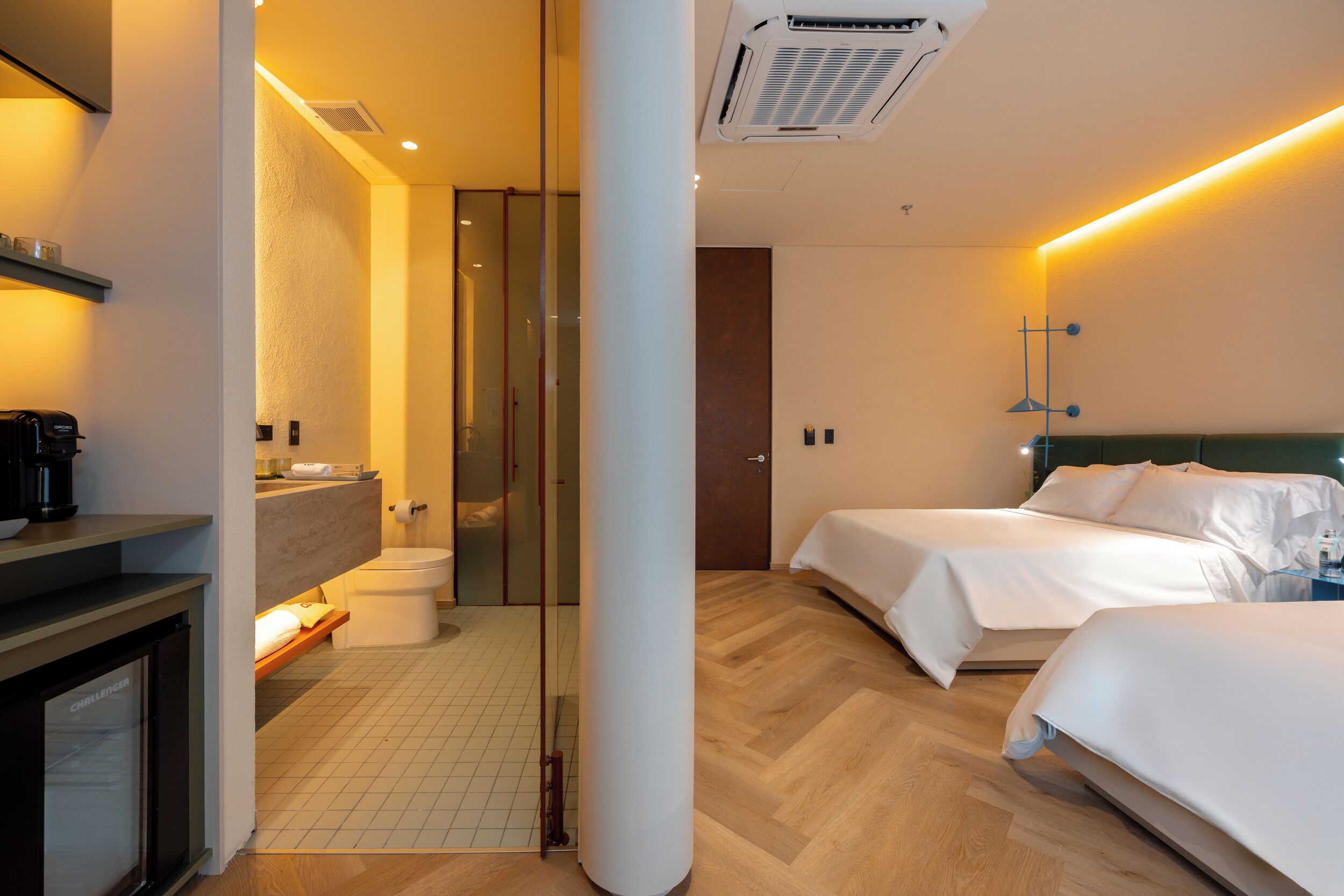 Quarto de hotel com banheiro, cama, mesa de cabeceira e criado-mudo.