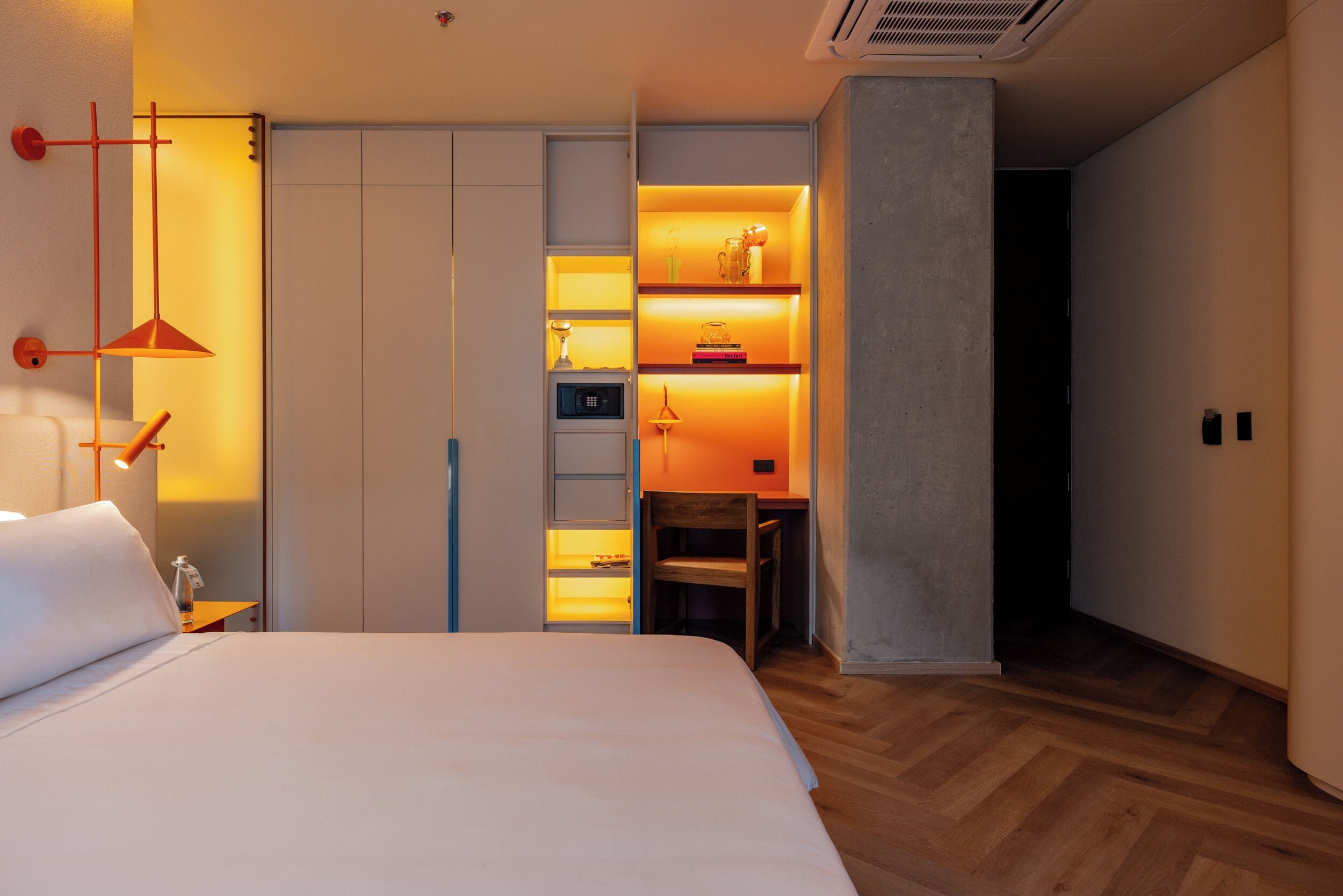 Ett modernt hotellrum med en stor säng, ett skrivbord och en inbyggd hyllsektion med böcker och dekorativa föremål.
