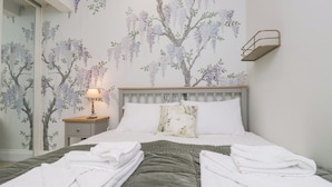 Un lit bien fait, avec du linge de lit blanc et une couverture verte, une table de chevet avec une lampe, et un mur orné d’un papier peint à motifs floraux.