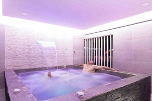 Een man ontspant in een bubbelbad met een waterval, omgeven door stenen muren en een paars verlicht plafond.