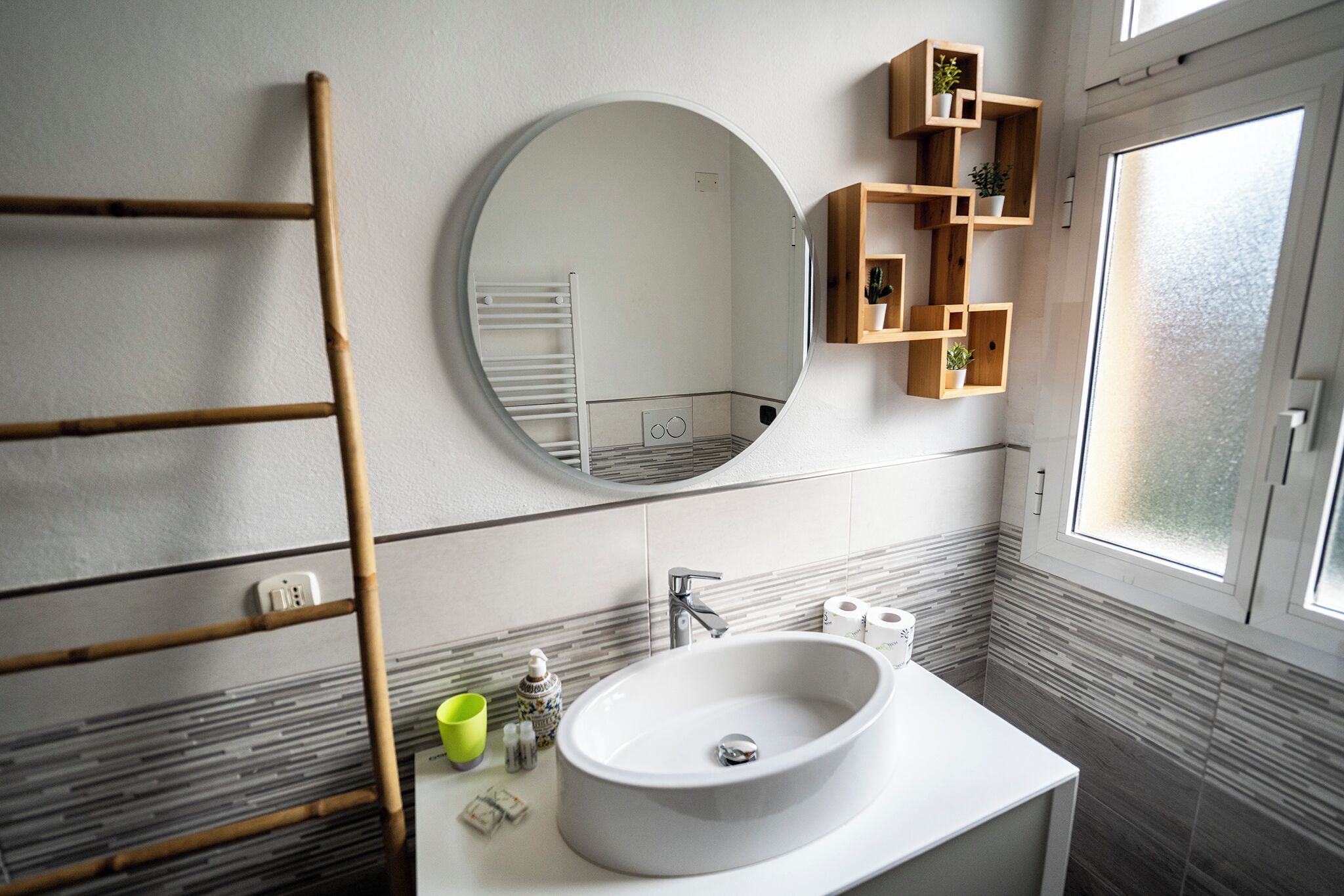 A bathroom with a round mirror, a white sink, a bamboo ladder, and a window with a view.