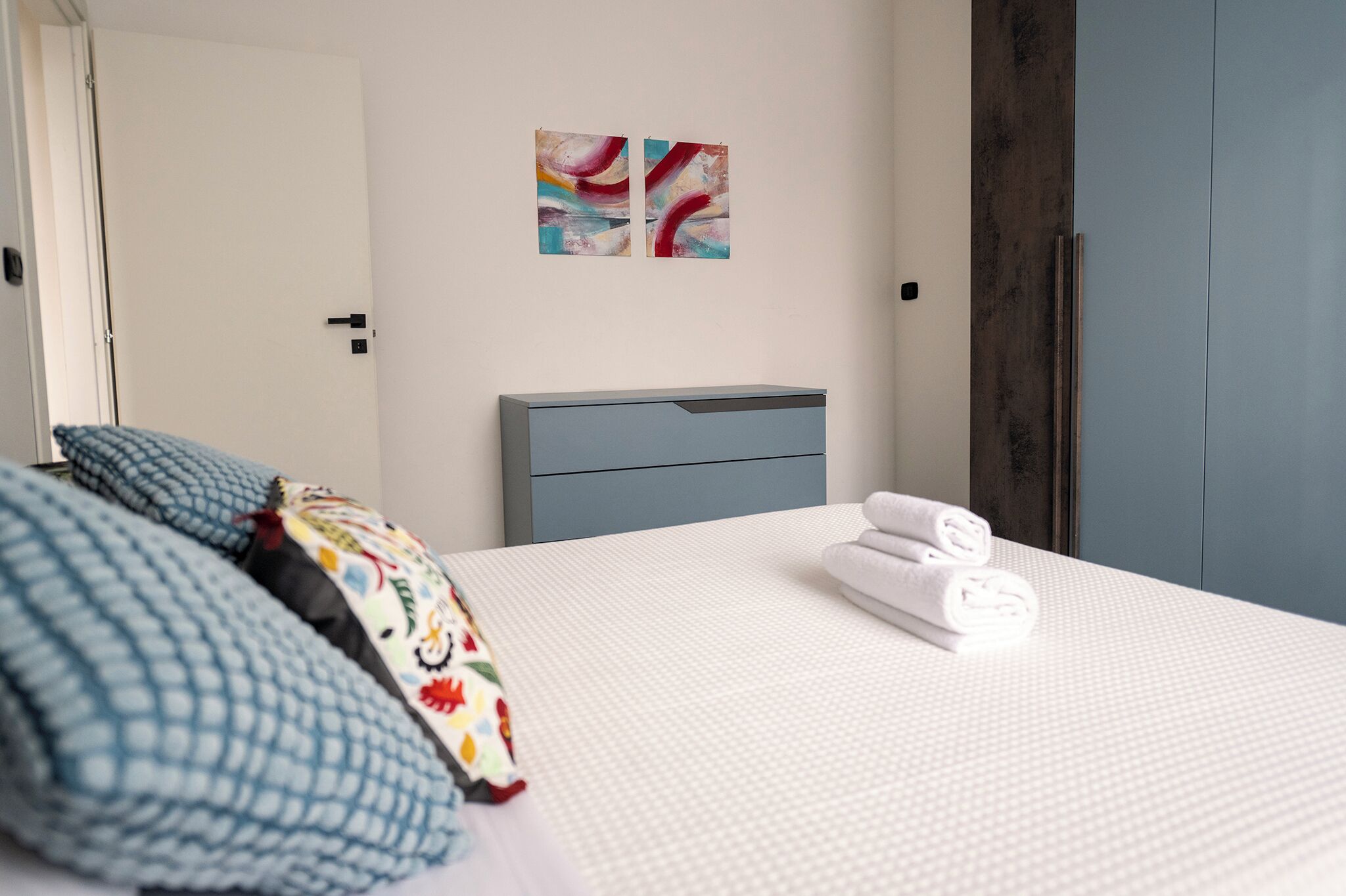 A hotel room with a bed, pillows, a headboard, a wall art piece, and a door.