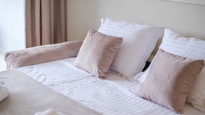 A neatly made bed with white linens and beige pillows, a rolled white towel, and a window with curtains.