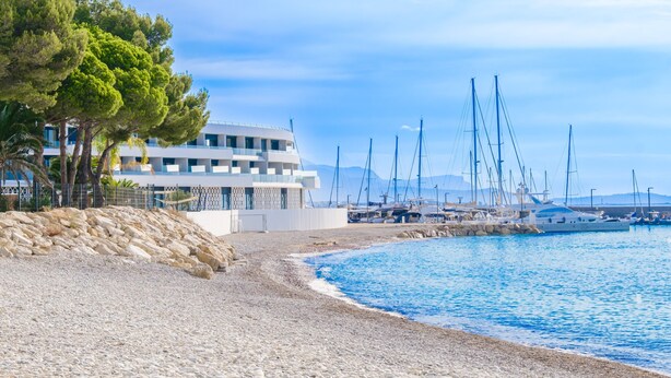 A coastal building with a rocky beach, sailboats, and a clear sky.