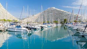 A marina with multiple boats docked, a large hotel with a unique curved architecture, and clear blue skies.