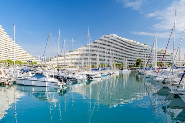 A marina with multiple boats docked, a large hotel with a unique curved architecture, and clear blue skies.