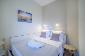 A neatly made bed with pillows and a folded towel, a small wooden side table, and a painting on the wall.