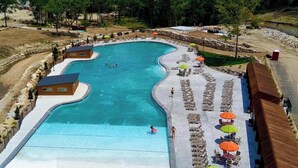 Une grande piscine incurvée, dotée de plusieurs espaces de détente et de parasols, entourée d’un mur de pierre et d’arbres.
