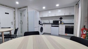 Une cuisine moderne avec des meubles blancs, un four à micro-ondes, un lave-vaisselle et un lave-linge. On y trouve une table à manger avec des chaises et une porte donnant sur l’extérieur.