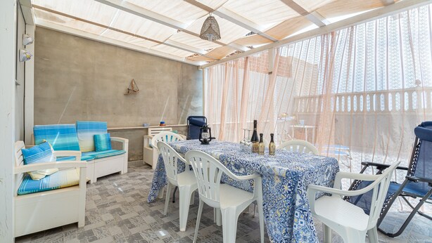 Un patio techado con una mesa lista para comer, sillas de plástico blanco y cojines con estampado azul en un sofá.