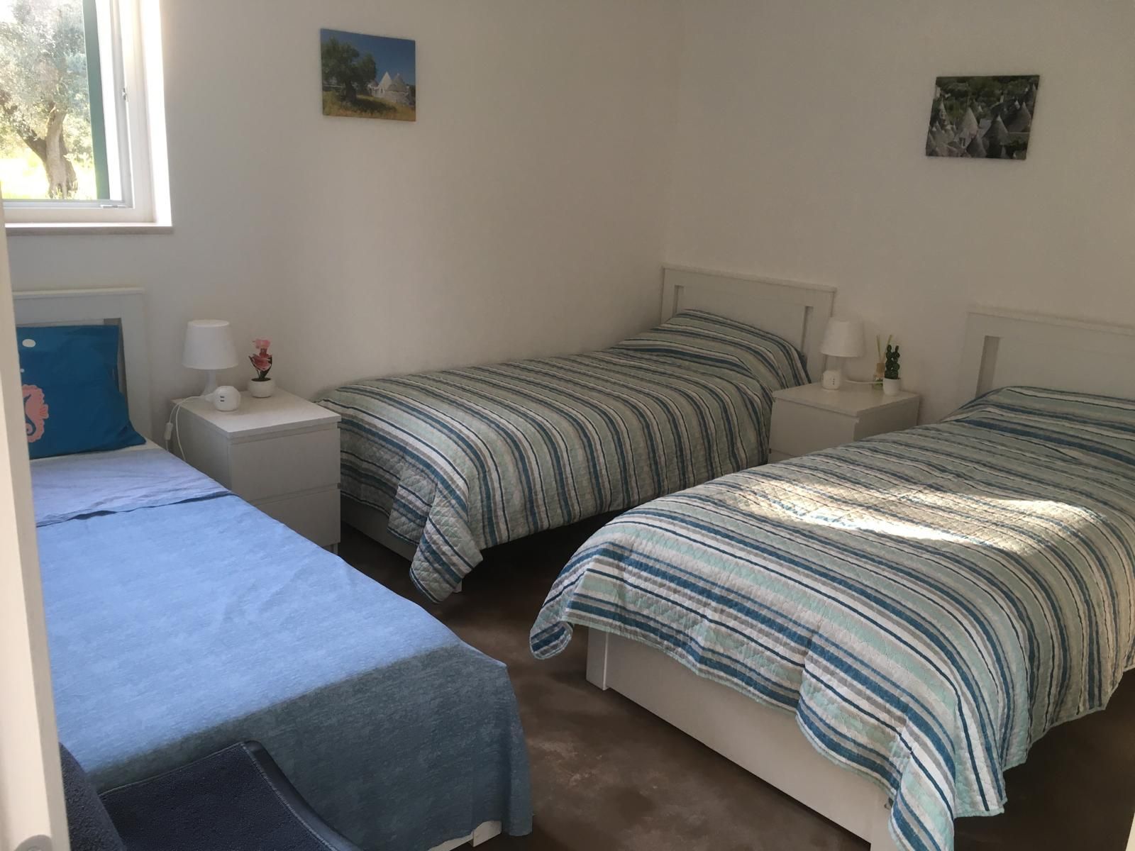 Una habitación con dos camas individuales, una mesita de noche con una lámpara, una pequeña mesa con un florero y una ventana con vistas a árboles.