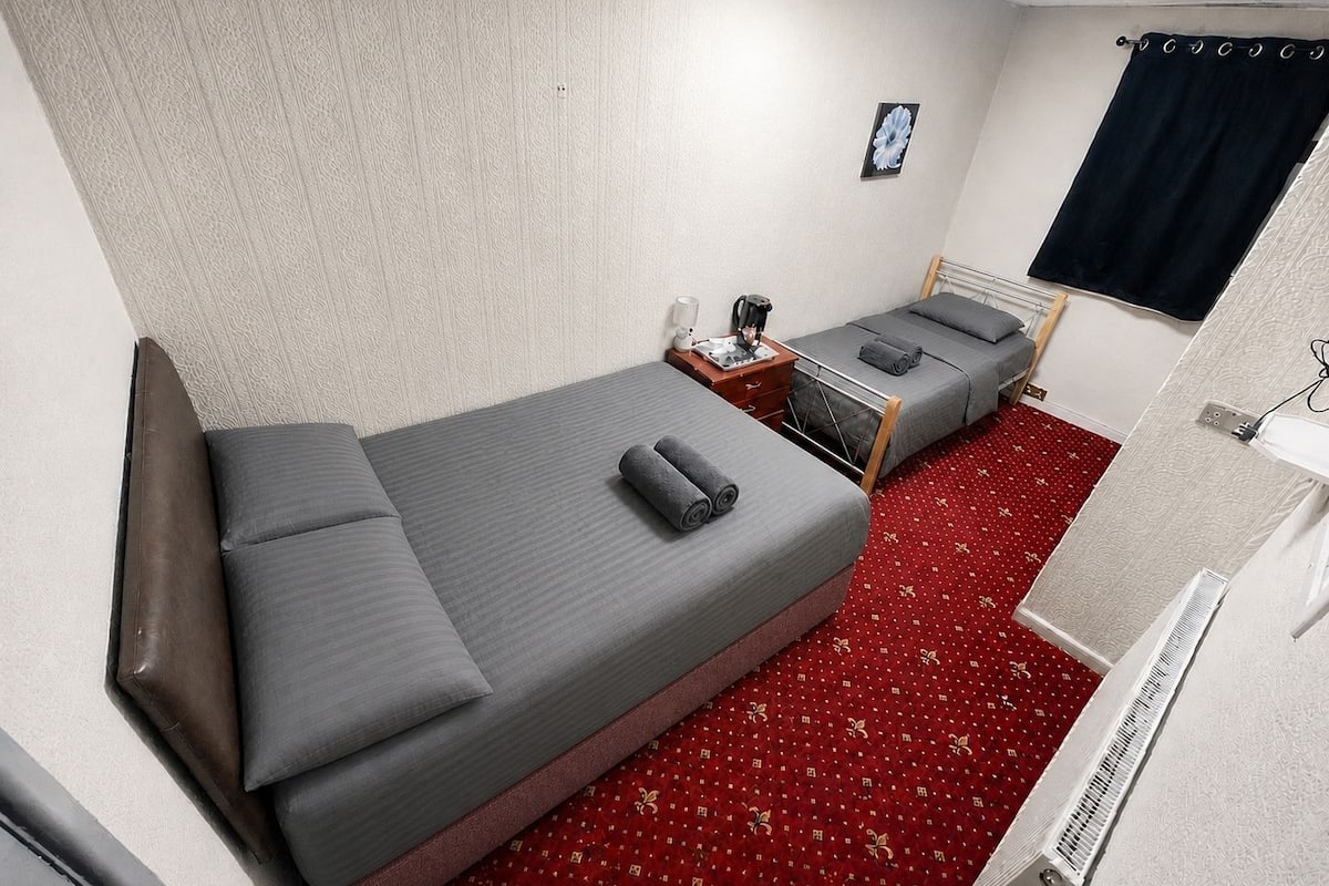 A single-bed room with a red carpet, a small bedside table with a lamp, a power outlet, and a window with a black curtain.