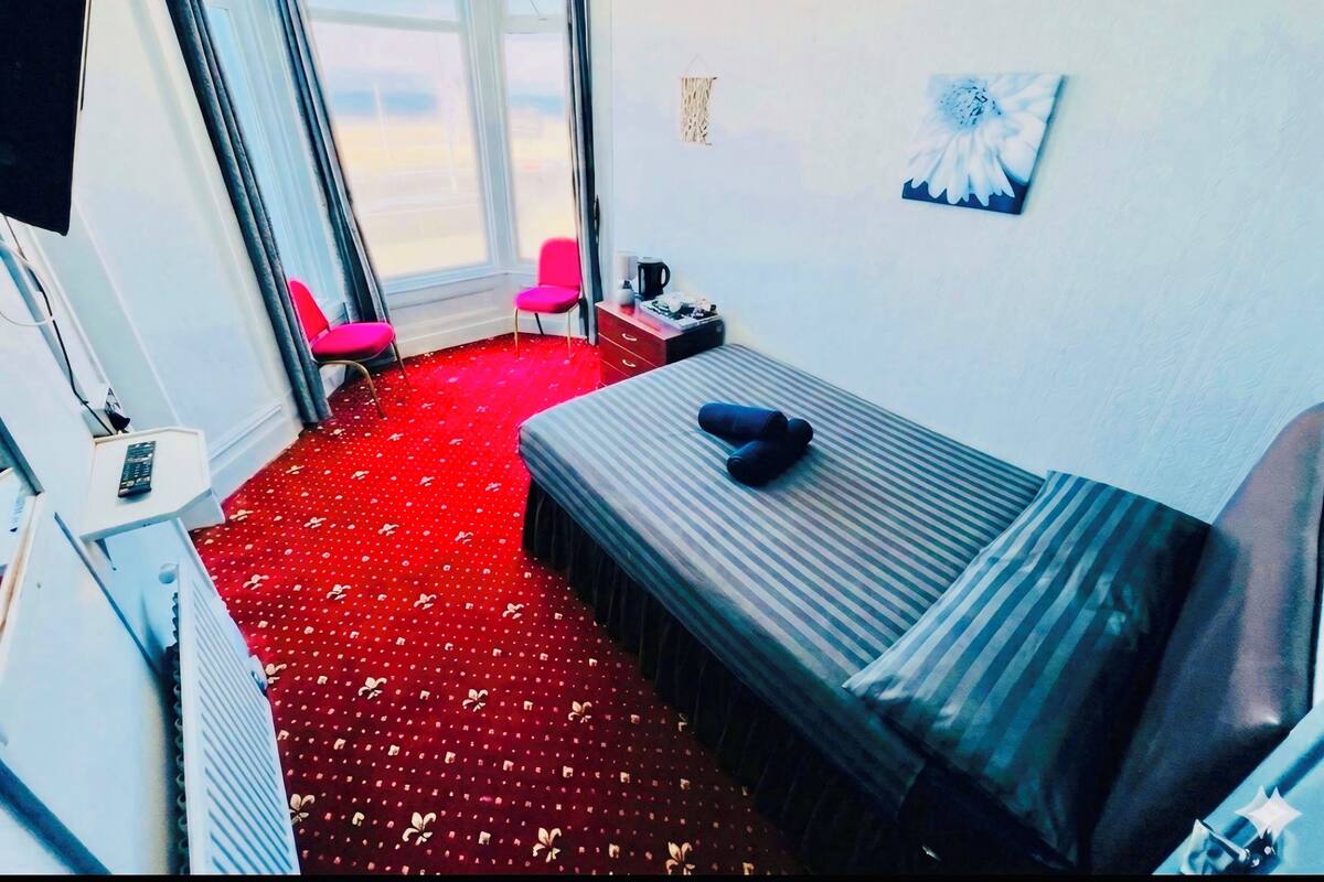 A hotel room with a bed, a chair, a desk with a computer, a window with curtains, and a painting on the wall.
