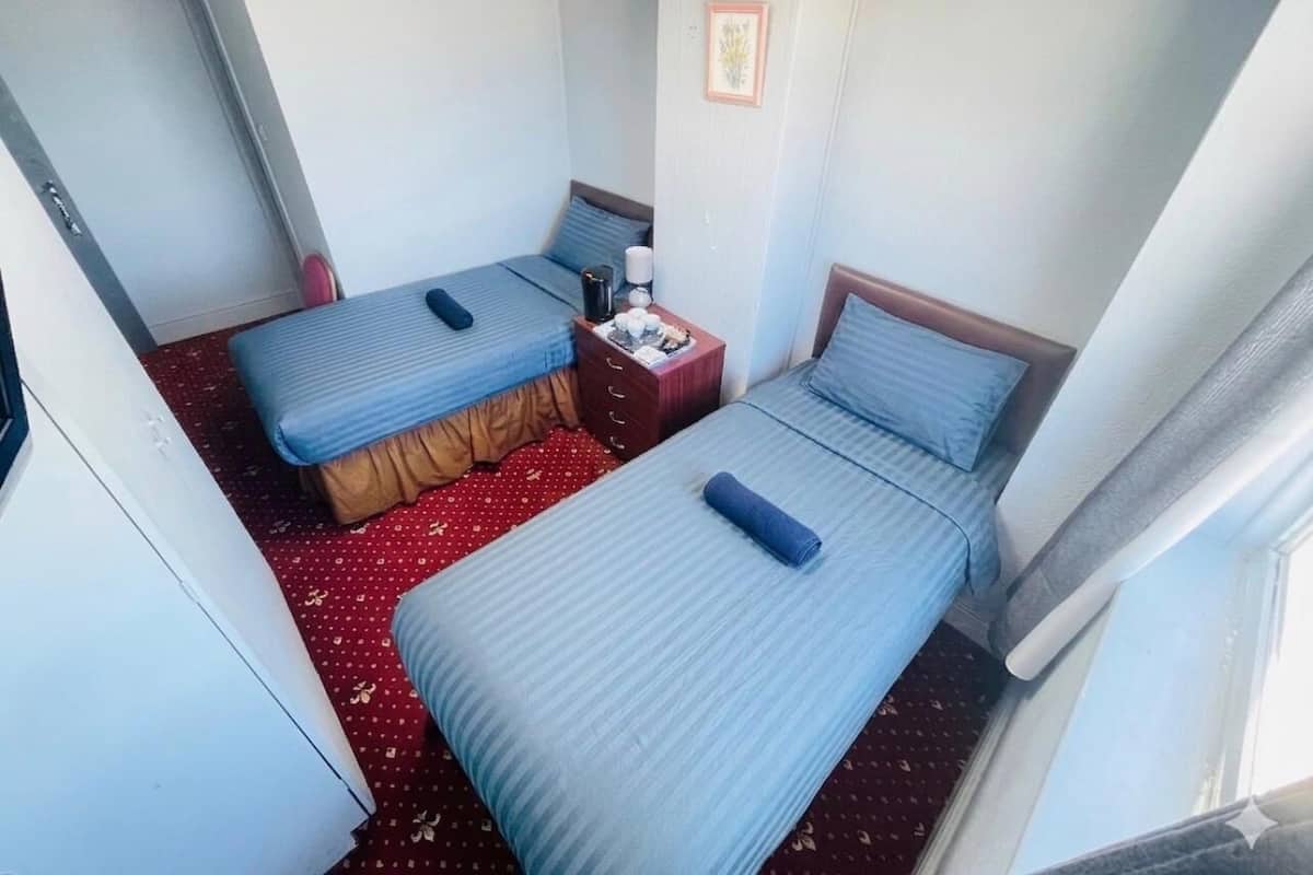 A hotel room with two beds, a small table with a lamp, and a window.