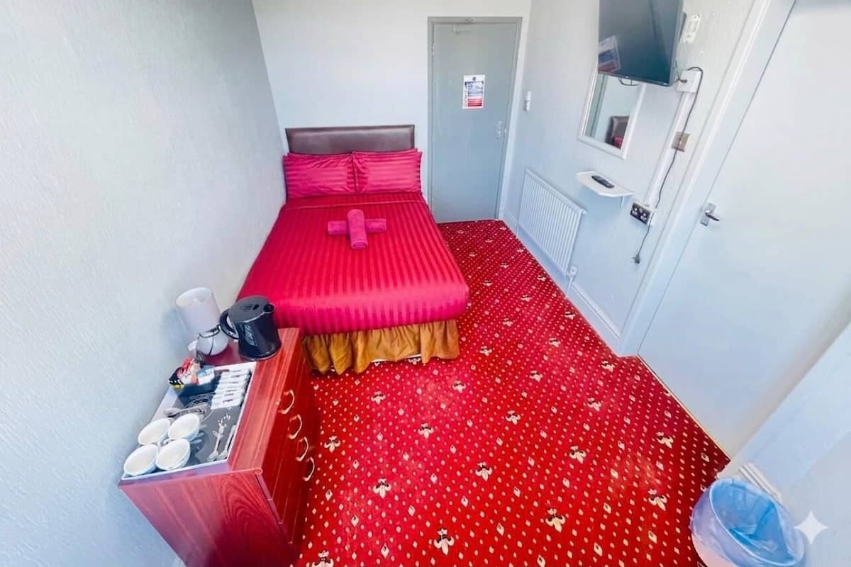 A small hotel room with a single bed, a TV mounted on the wall, a small table with a tray of items, and a red patterned carpet.