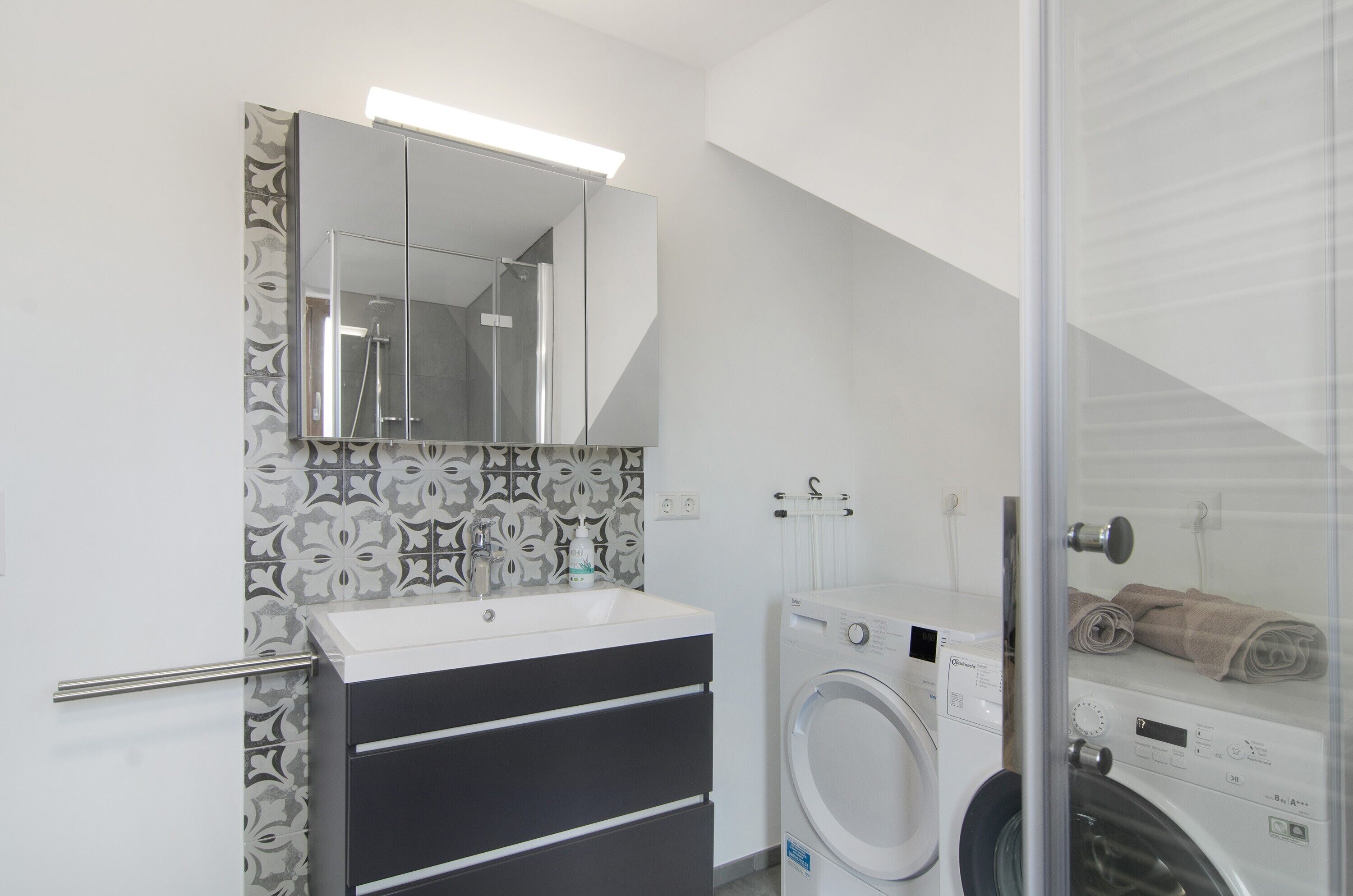 Uma casa de banho moderna com um espelho grande, uma pia, uma máquina de lavar roupa e um chuveiro com cabine de vidro.