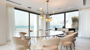A modern dining room with a round glass table surrounded by beige chairs, large windows with a view of the sea, and a contemporary chandelier.