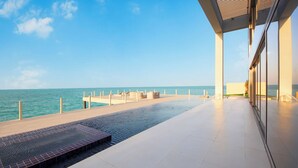 Una piscina infinita moderna con vista al océano, un área techada con asientos y un cielo despejado.