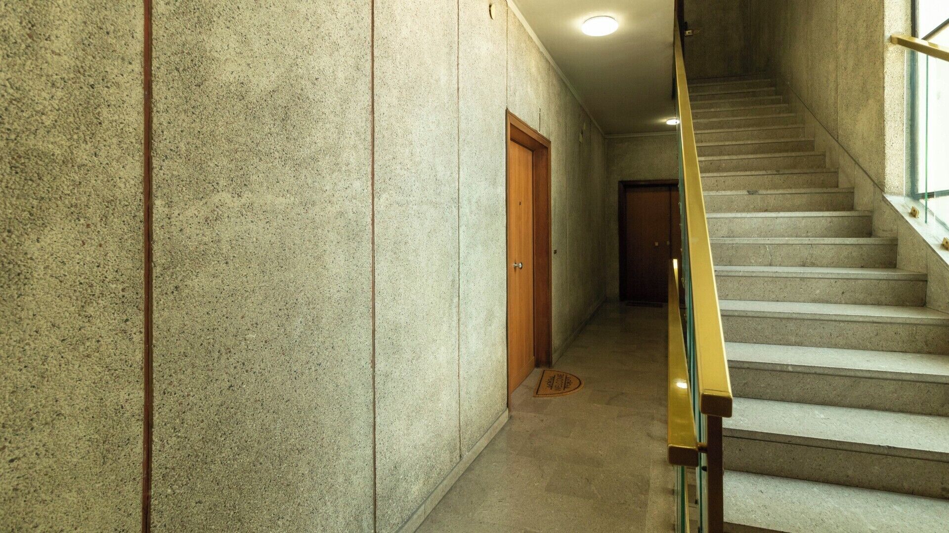 A hallway with a concrete wall, a wooden door, and a staircase with a metal handrail.