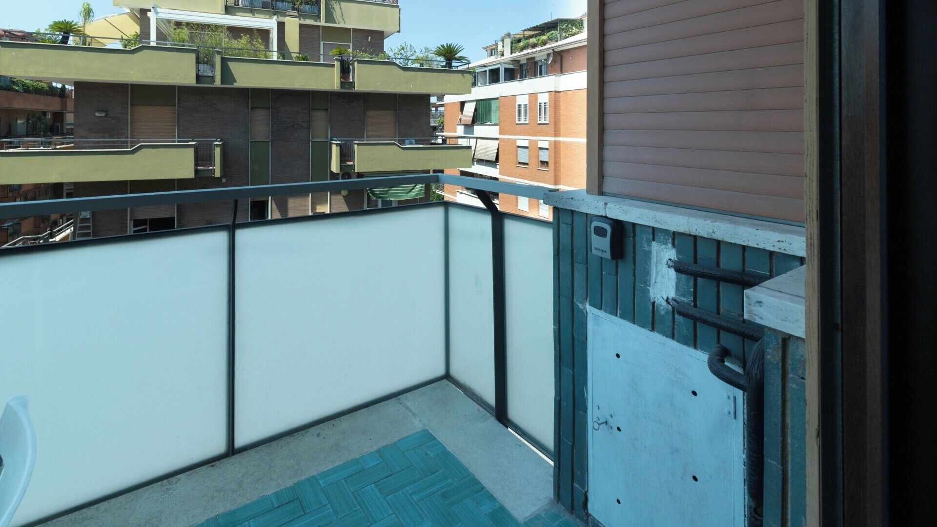 Un balcon avec un tapis bleu, une rambarde en métal et une vue sur les bâtiments voisins.