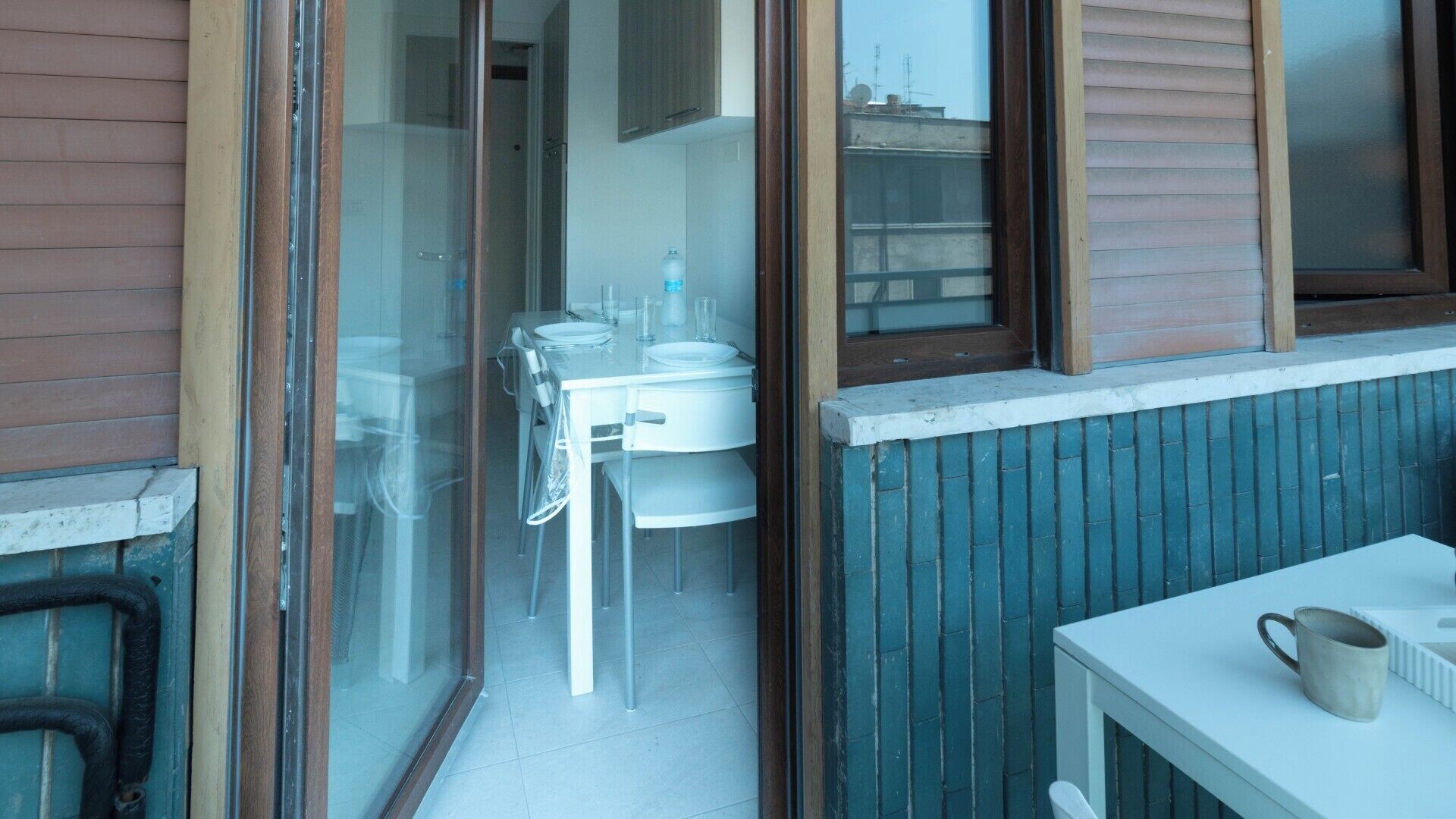 Une porte vitrée donne sur un coin repas, où se trouvent une table et des chaises, une fenêtre avec des stores et une petite table sur laquelle est posée une tasse, sur la droite.
