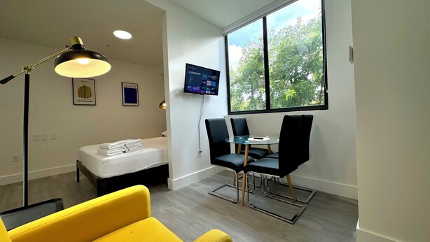 Une chambre moderne avec un lit, un fauteuil jaune, une table à manger avec des chaises, un téléviseur fixé au mur et une grande fenêtre donnant sur des arbres.