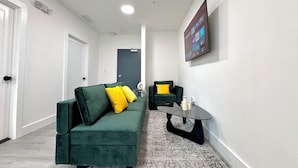Un salon moderne avec un canapé vert, des coussins jaunes, une petite table ronde et un téléviseur à écran plat fixé au mur.