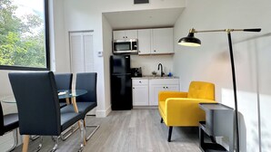 Une cuisine moderne avec des armoires blanches, un réfrigérateur noir et un four à micro-ondes. On y trouve un coin repas avec une table en verre et des chaises noires. Un fauteuil jaune est visible au premier plan.