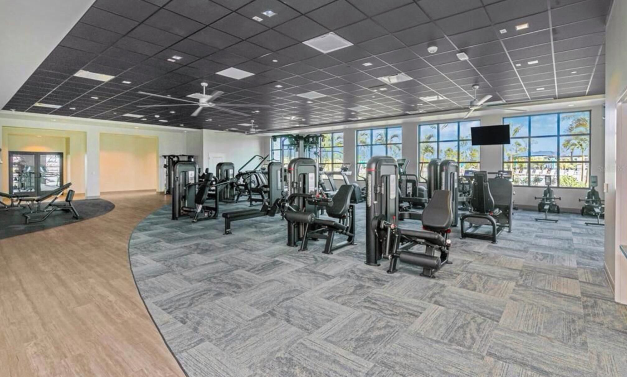 Un gimnasio con diversas máquinas de ejercicio, ventanas amplias y un ventilador de techo.