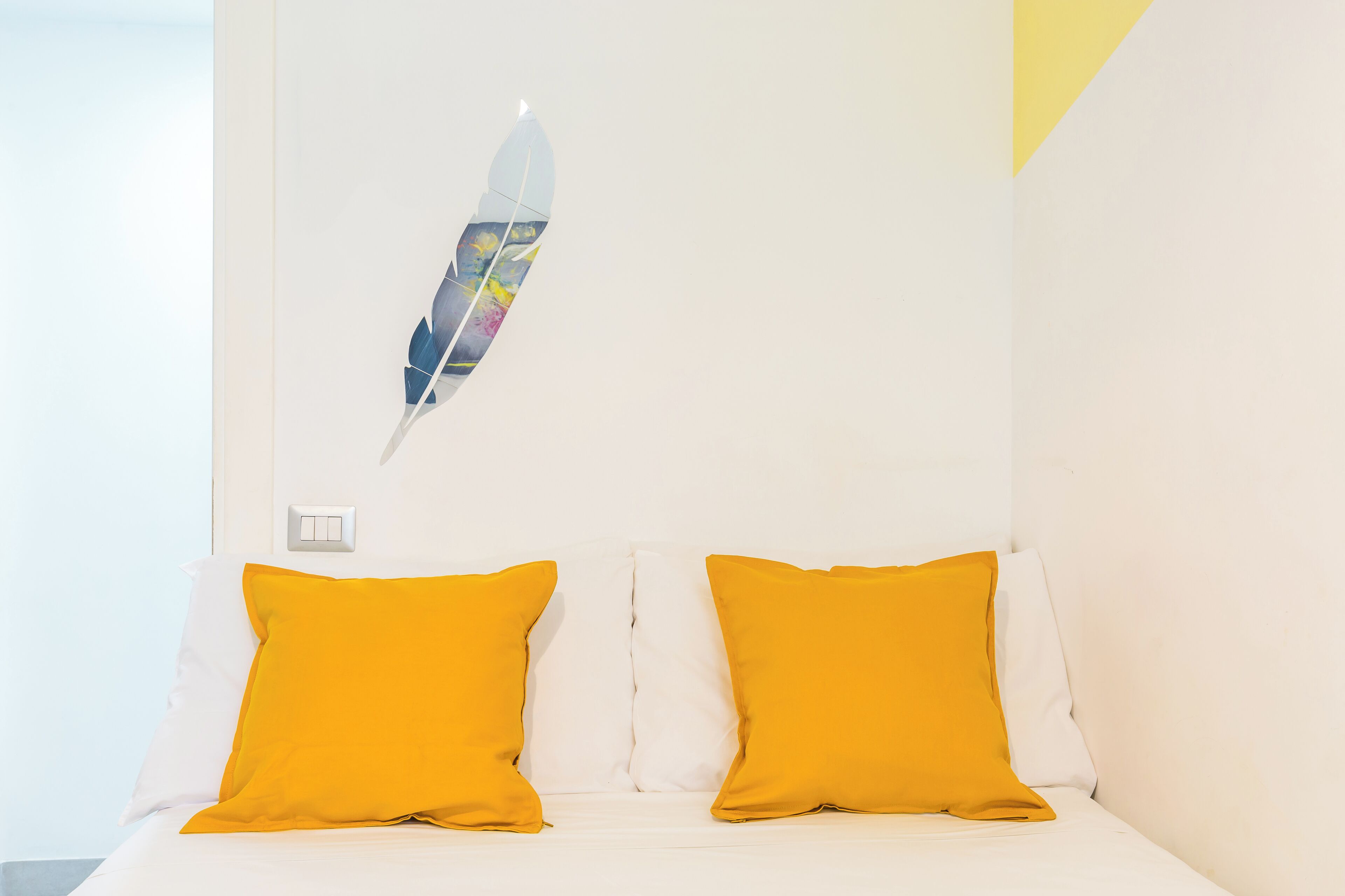 A bed with white bedding and two bright yellow pillows, a white wall, and a single white light switch.
