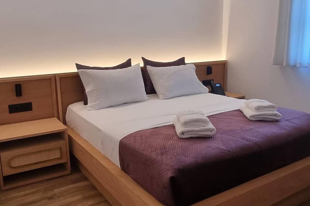 Une chambre d’hôtel moderne avec un cadre de lit en bois, du linge de lit blanc et un plaid violet. On y trouve une table de chevet en bois avec une lampe et un petit tiroir.
