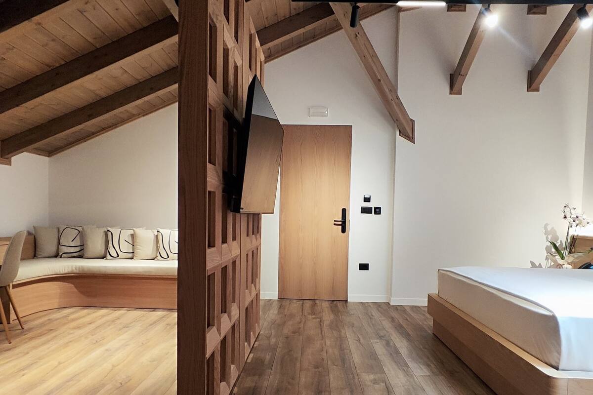 Une chambre spacieuse située dans les combles, avec des poutres apparentes, un lit en mezzanine, une banquette intégrée avec des coussins et une armoire en bois.