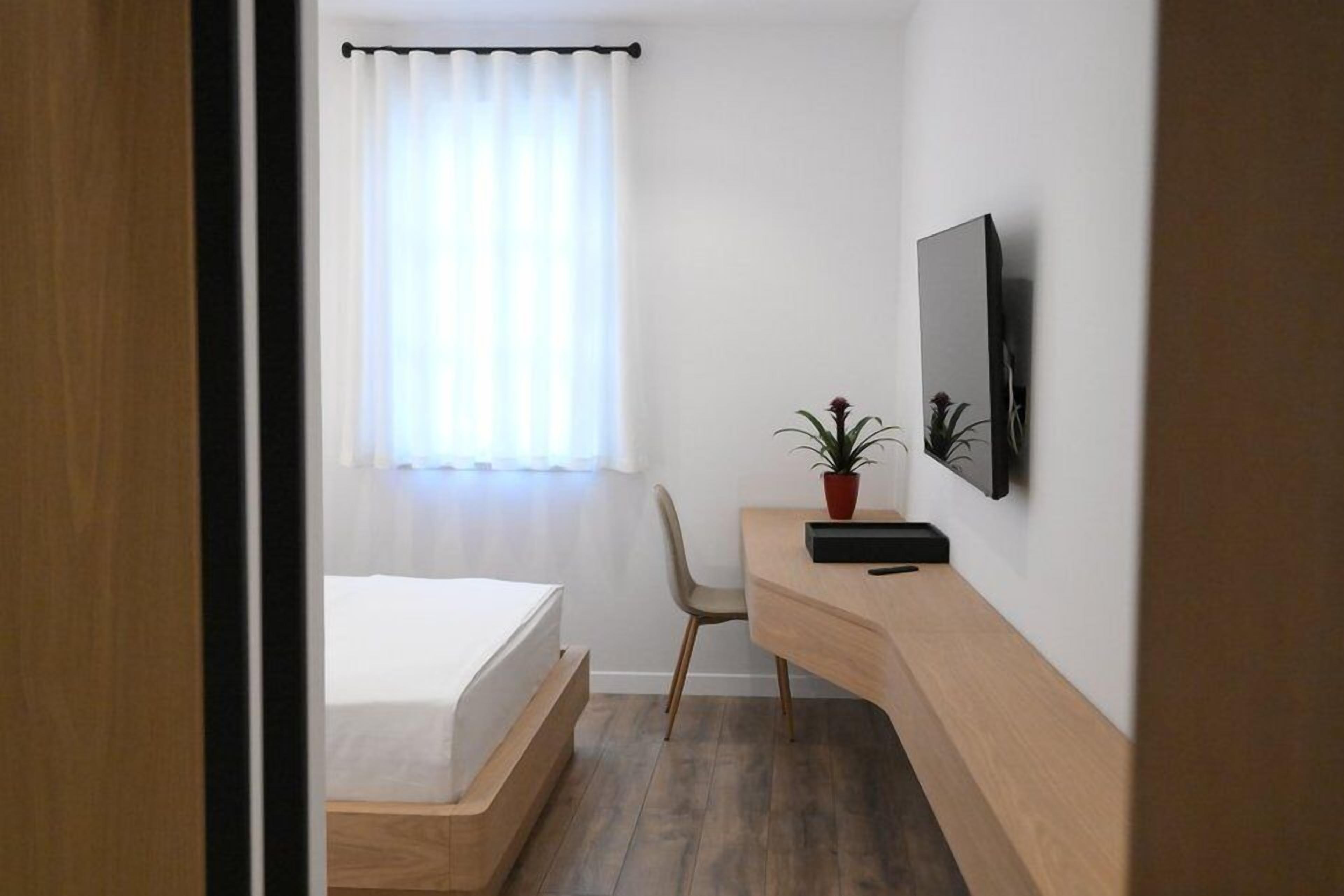 A modern hotel room with a bed, desk, chair, potted plant, and a mounted TV.