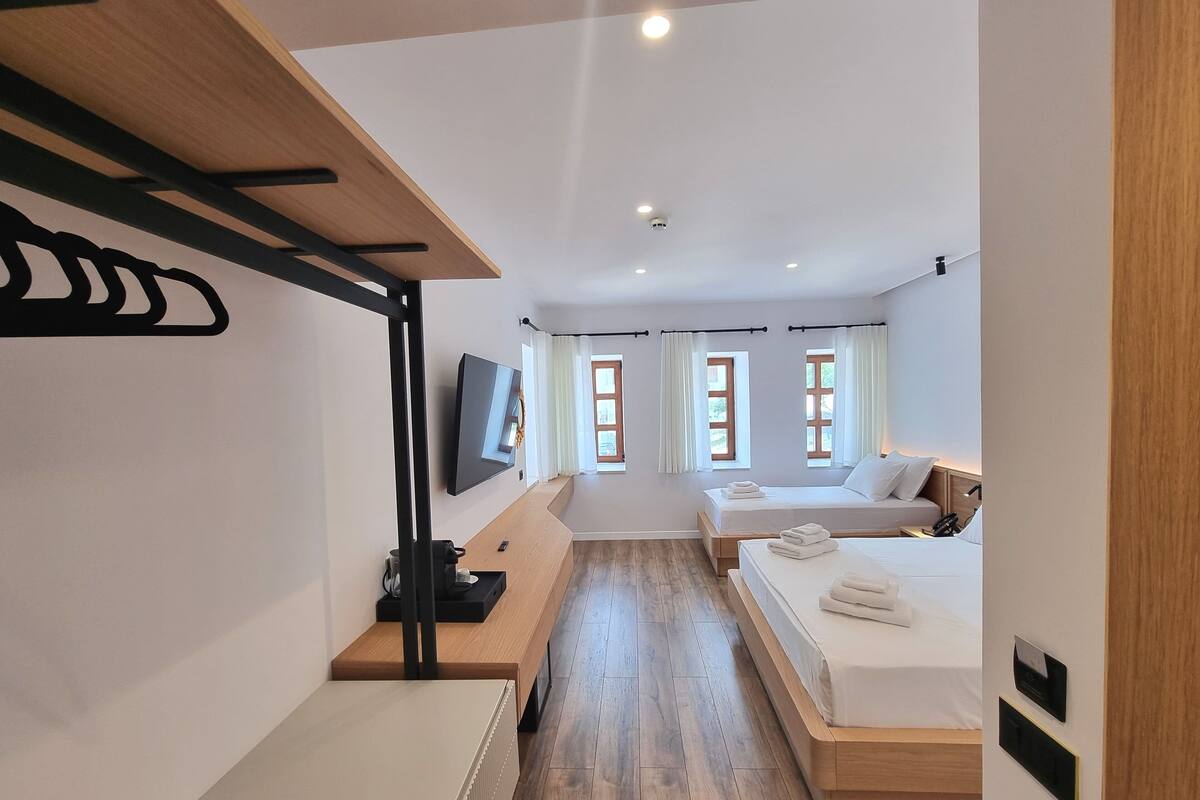 Une chambre d’hôtel moderne avec un plancher en bois, un lit recouvert de draps blancs, un bureau avec un ordinateur, un téléviseur fixé au mur et une fenêtre avec des rideaux.