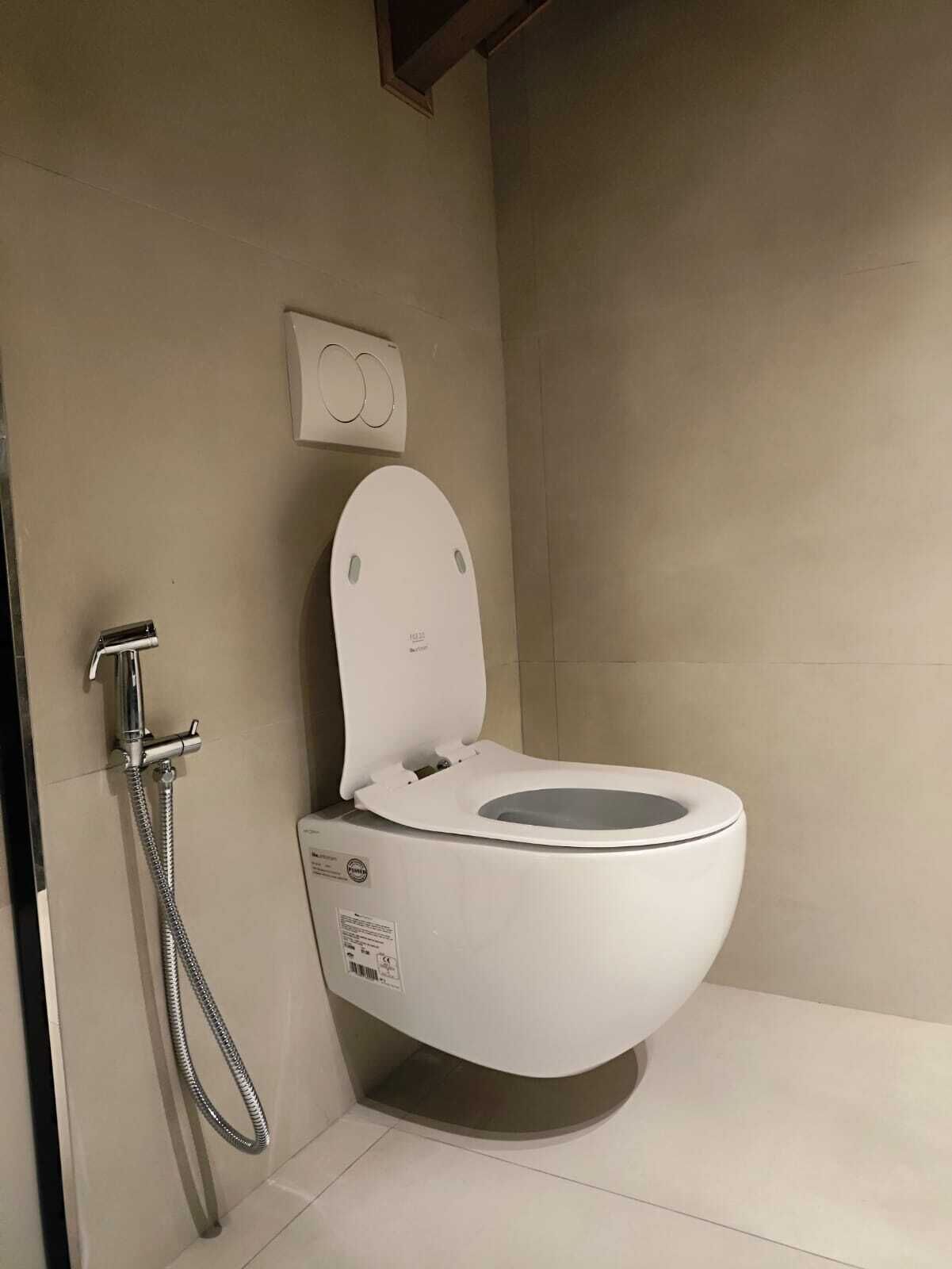 Un baño moderno con inodoro blanco, cabezal de ducha de mano y botón de descarga en la pared.