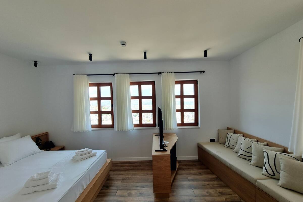 Une chambre à coucher avec un lit, un canapé, deux fenêtres avec des rideaux, une télévision et un plancher en bois.