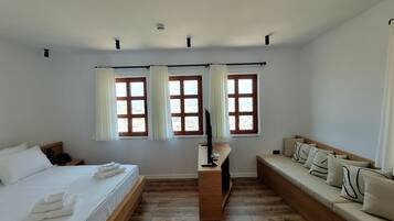 A bedroom with a bed, a sofa, two windows with curtains, a TV, and a wooden floor.