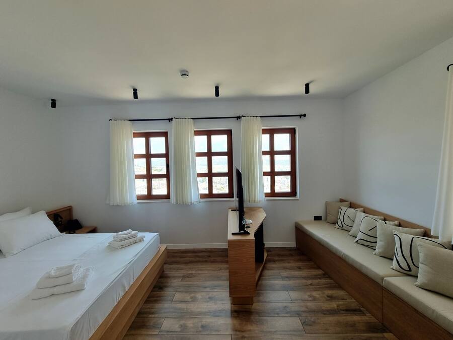 A bedroom with a bed, a sofa, two windows with curtains, a TV, and a wooden floor.